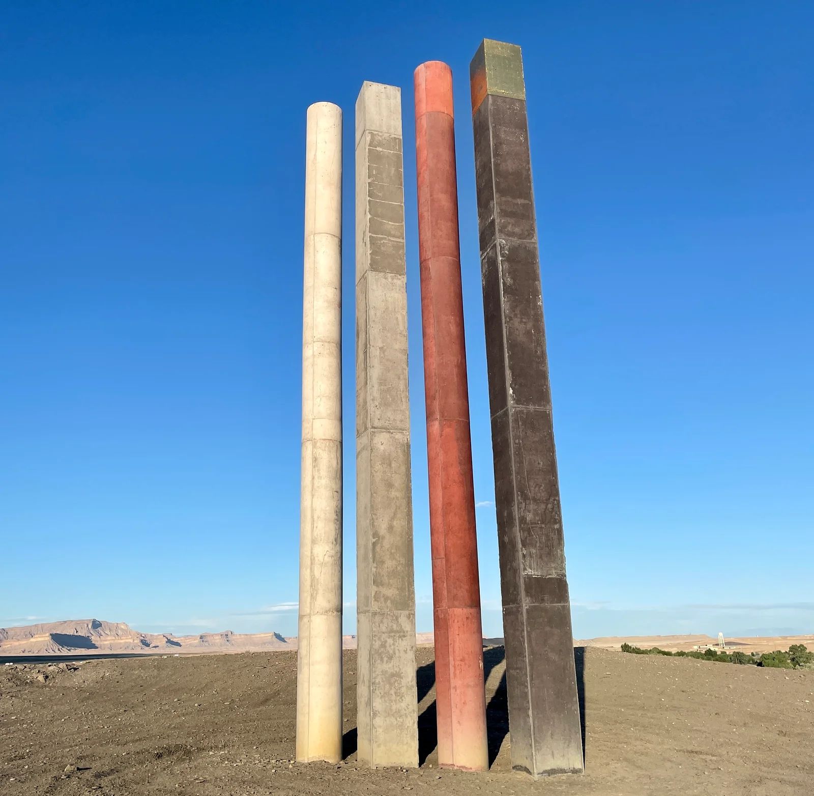 Five tall, rectangular, differently colored concrete pillars stand on a desert landscape under a blue sky.