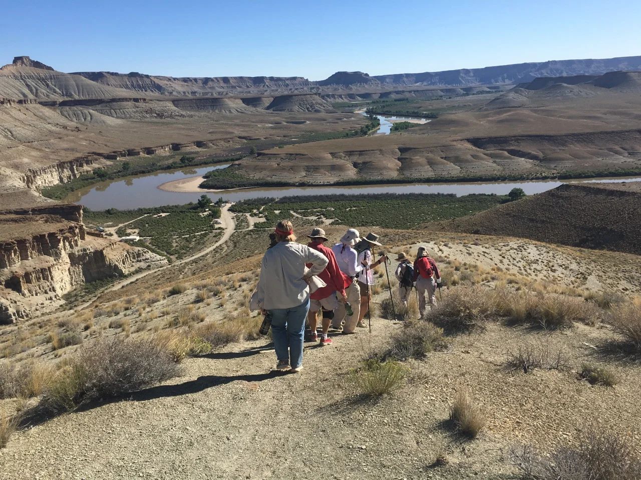 Hikers on a hillside overlook a river winding through a desert canyon under a bright sky.