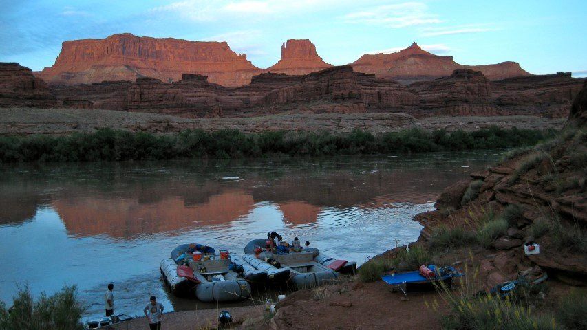 Rafts on a river at dusk, reflecting red rock cliffs. People on the shore.
