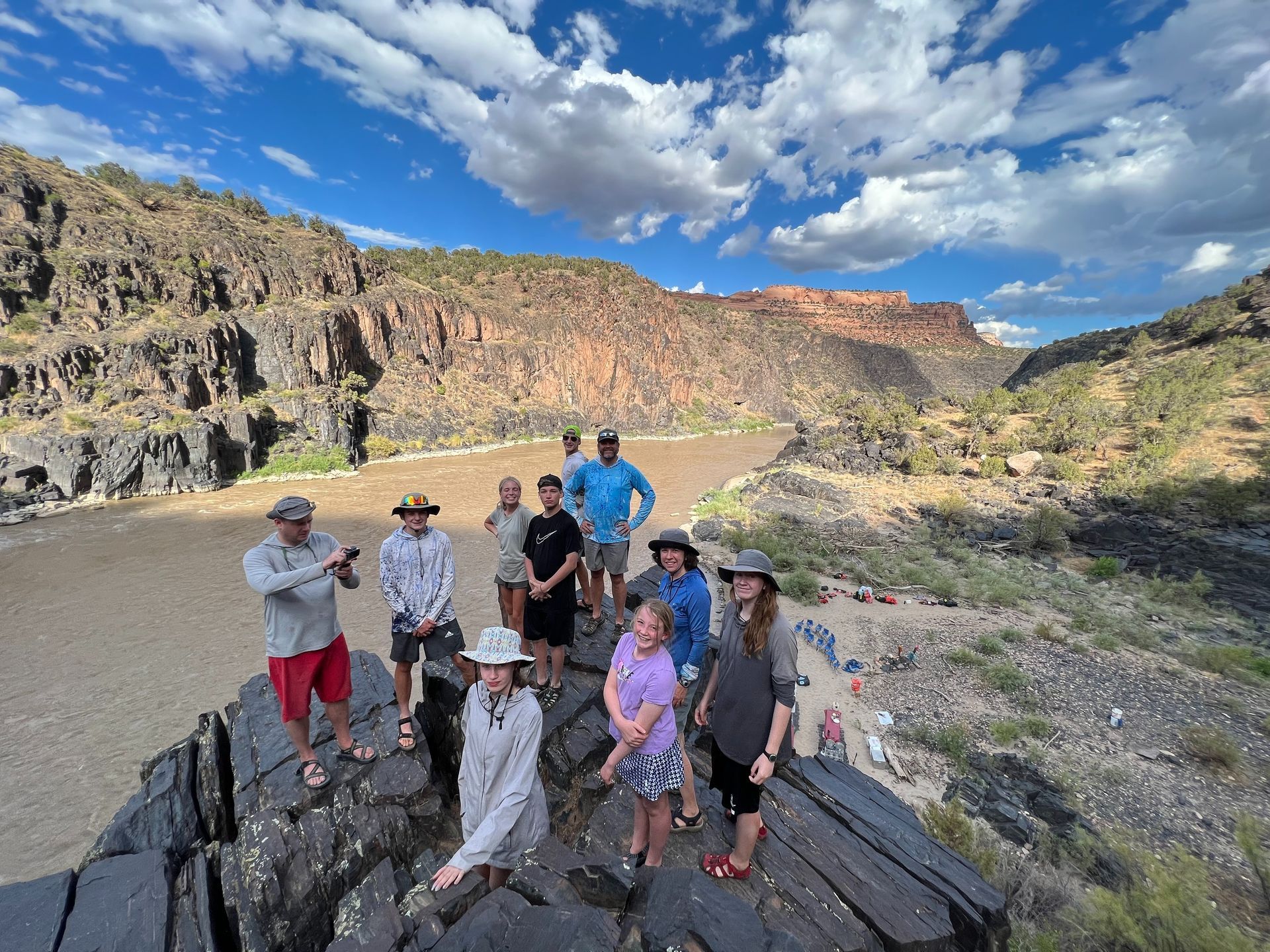 Group of people on a rocky outcrop, overlooking a canyon river. Blue sky, brown cliffs.