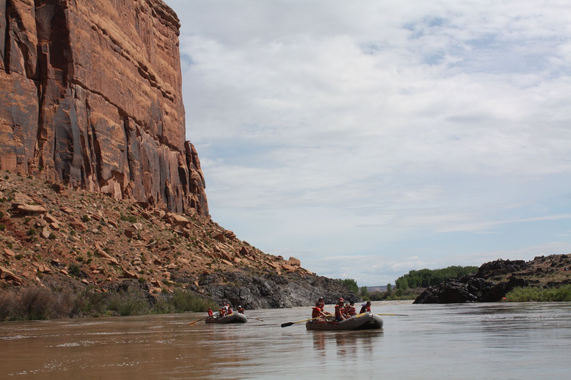 Rafters on a muddy river near a towering red rock cliff under a cloudy sky.