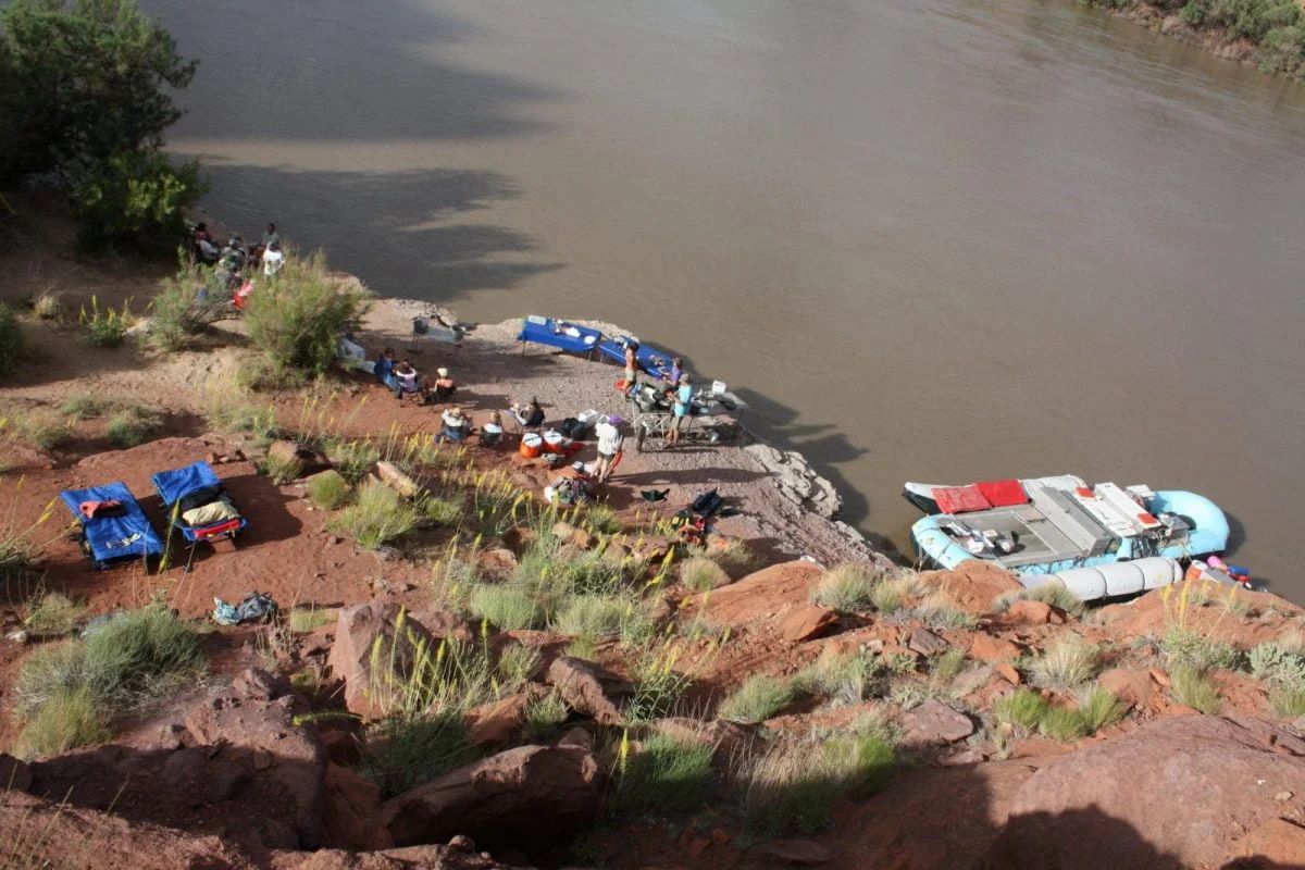 Rafts on muddy river bank with people, gear, and tents, in a canyon setting.