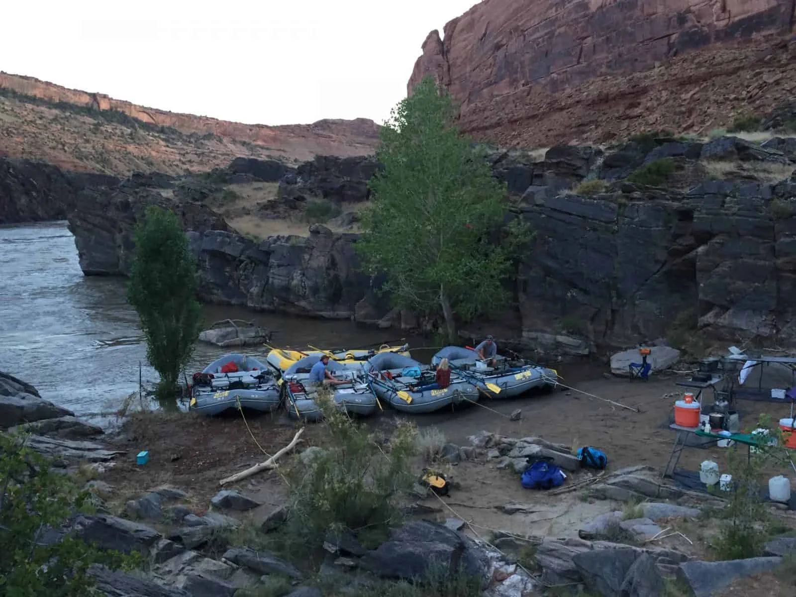 Rafts lined up on a rocky shore next to a river. Camping gear and trees are also present.