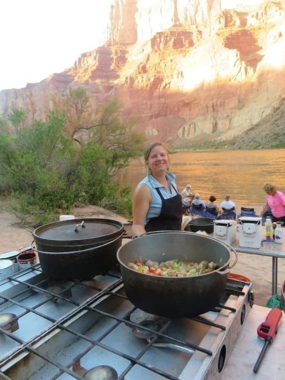 Woman cooking with Dutch ovens on a camping stove near a river. Canyon backdrop, sunny.