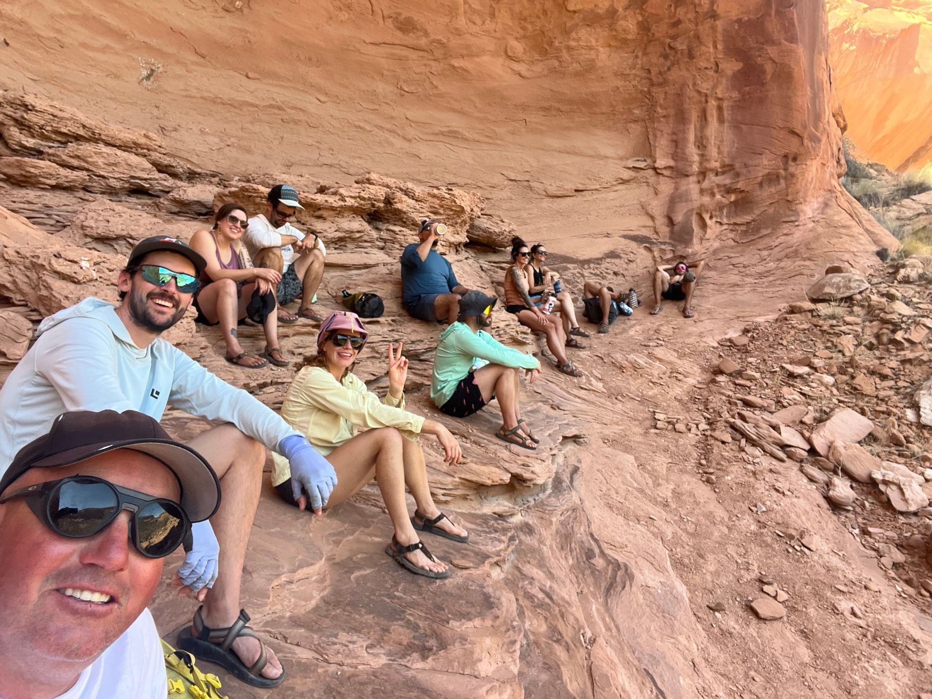 Group of people resting under a red rock overhang in a desert setting.