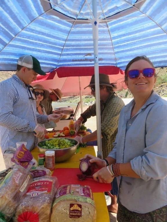 People preparing food at a picnic table under a striped umbrella. Woman smiles while chopping red onion.