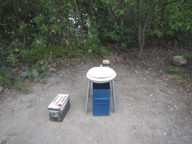 Outdoor portable toilet with a battery next to it, set in a wooded area.