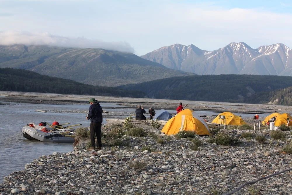Campsite along a riverbank with tents, raft, and mountains in background. People are gathered, enjoying the outdoors.