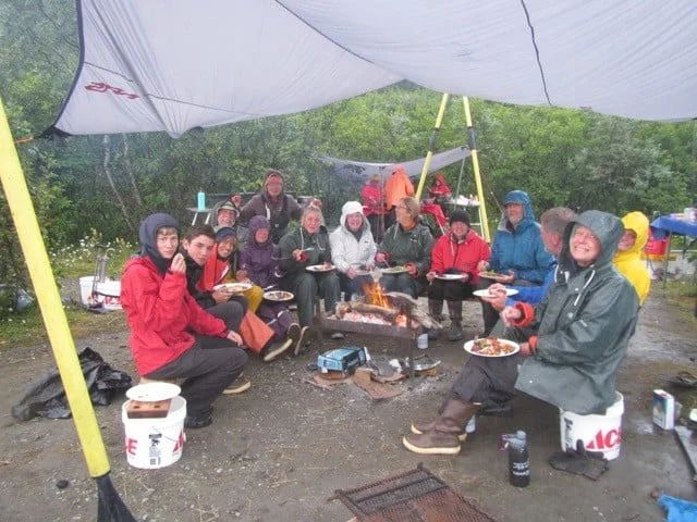 People gathered around a campfire under a tarp, eating. Some wear rain gear. Outdoors.