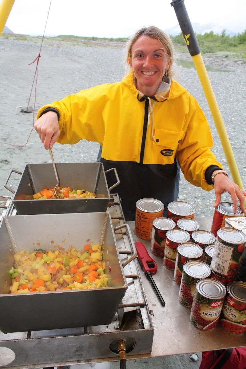 Woman in yellow raincoat cooks a meal outdoors, stirring food in metal pans; cans of food on a table.