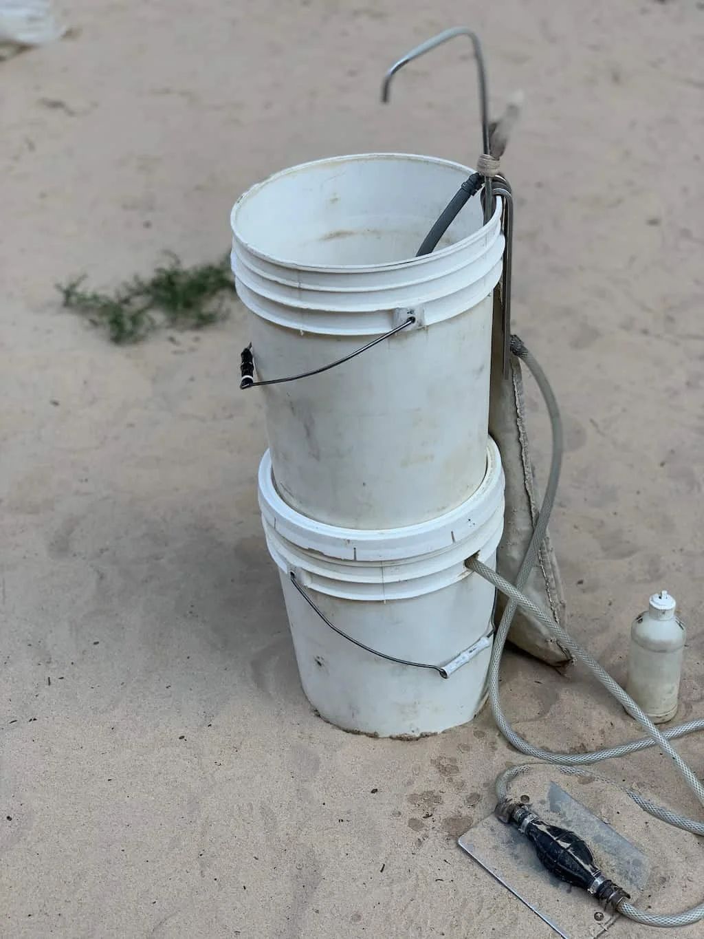 Two white buckets stacked, makeshift handwashing station with hose and tap on sand.