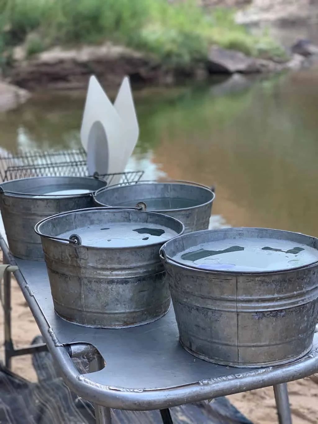 Four metal buckets filled with water sit on a table by a river.