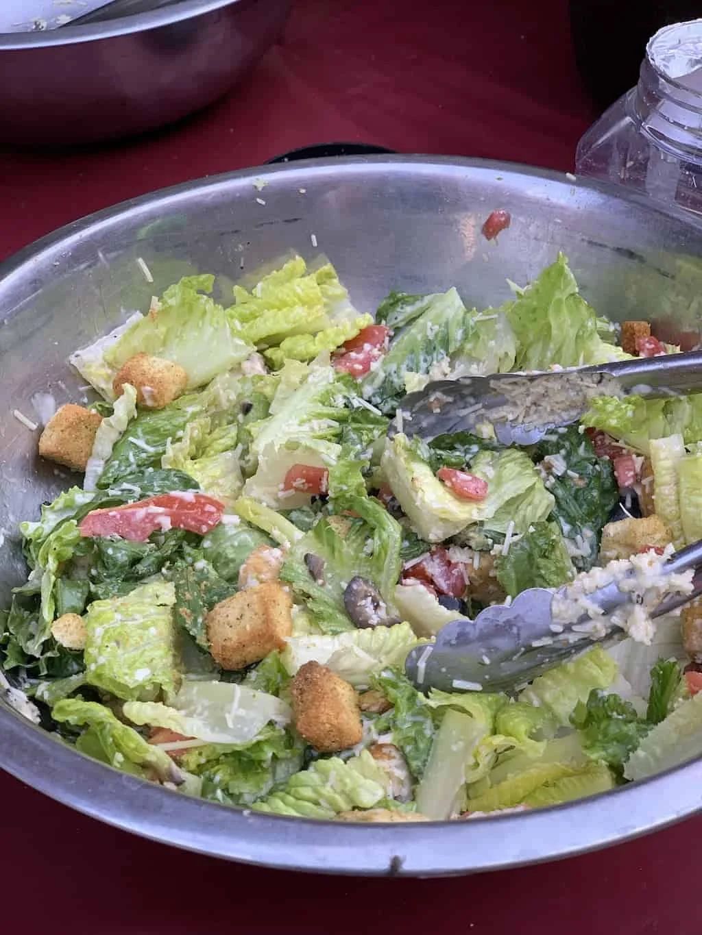 Caesar salad in a metal bowl with tongs, croutons, and tomatoes.