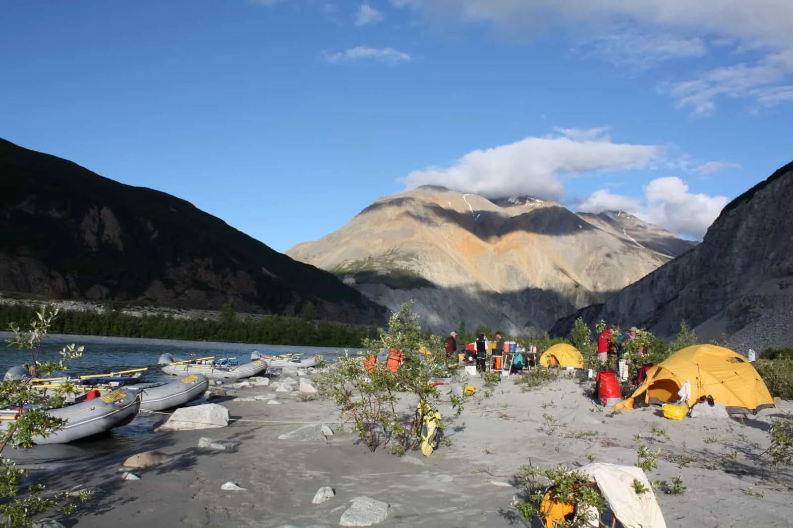 Campers near rafts and tents on a sandy shore with mountains and river in the background under a blue sky.