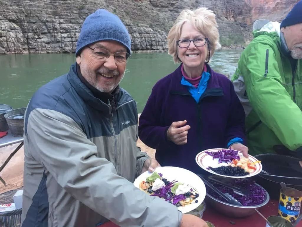 People holding plates of food near a river. Man wears a blue hat, smiling. Woman smiles, holding a plate of salad.