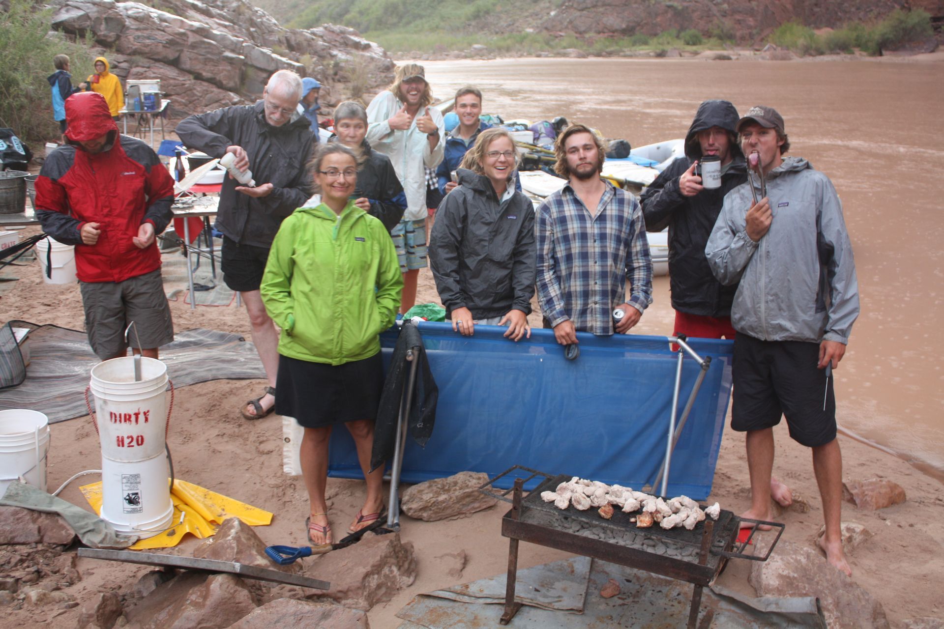 Group of people standing by a river, some grilling food, cloudy weather.