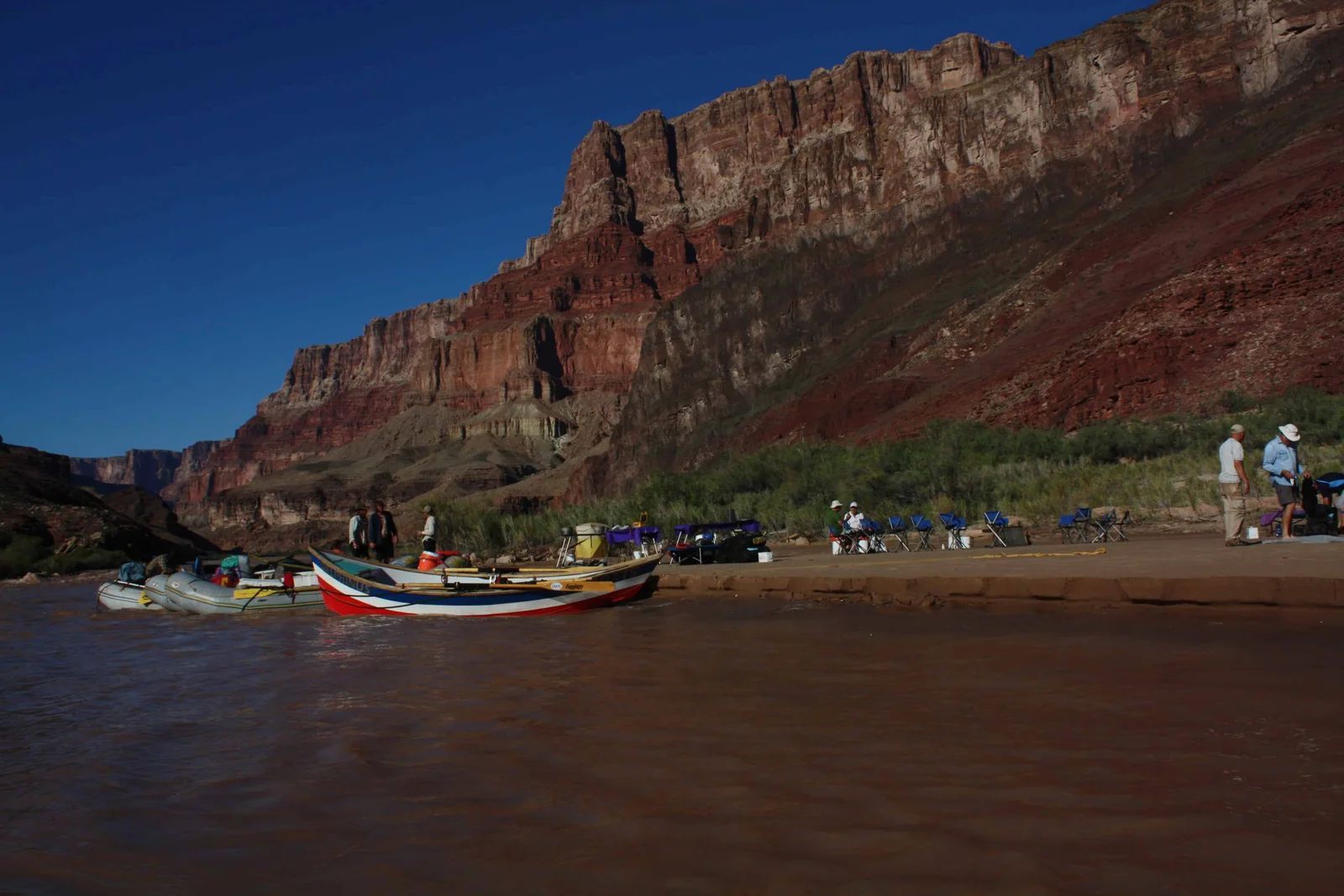 Boats and people on a riverbank next to a large red cliff under a blue sky.