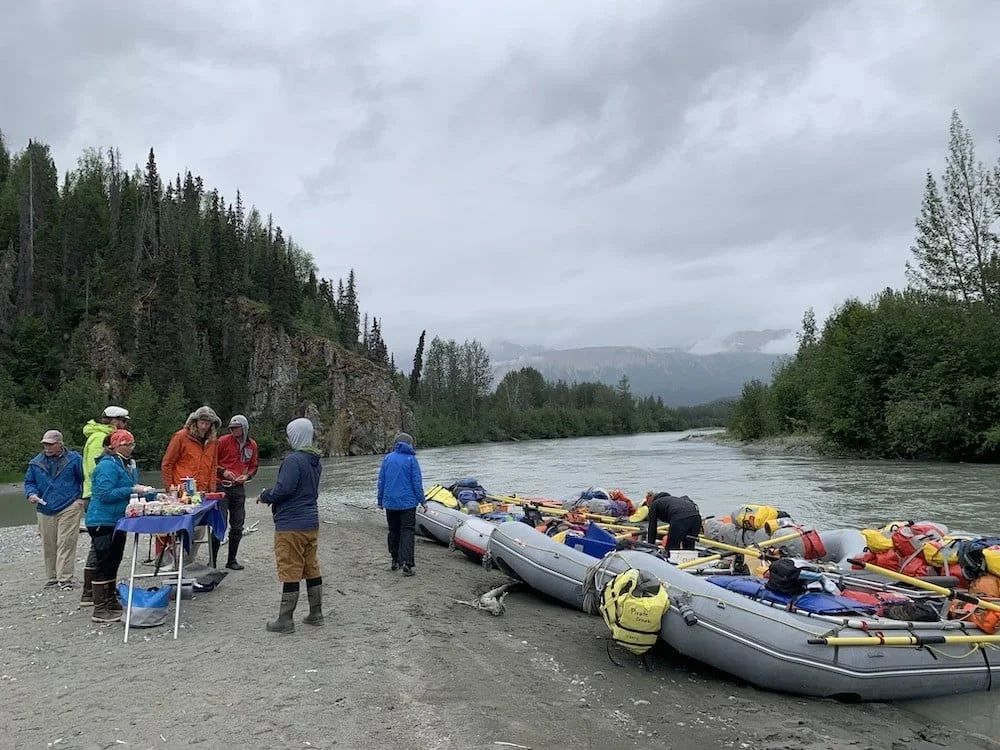 People near rafts on a riverbank, preparing for a rafting trip under an overcast sky with mountains in the distance.