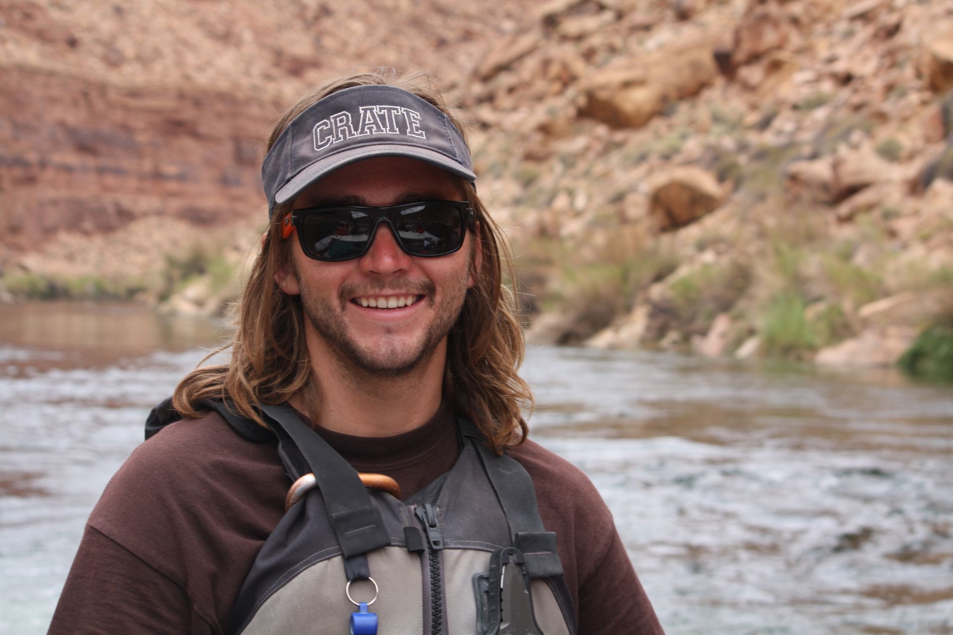 Man with long hair and sunglasses smiles by a river in a canyon. He wears a visor and life vest.