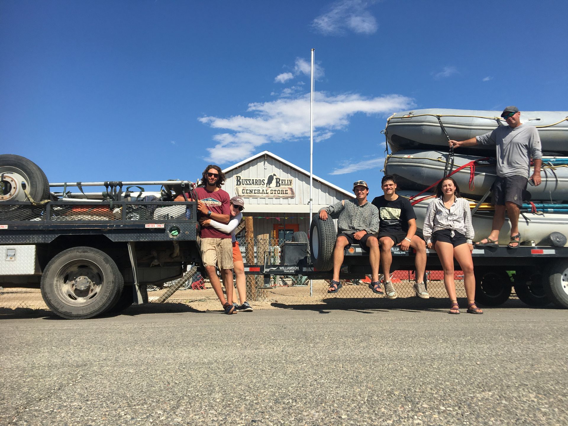 Group stands in front of a building with a truck loaded with rafts. Sunny day, blue sky.