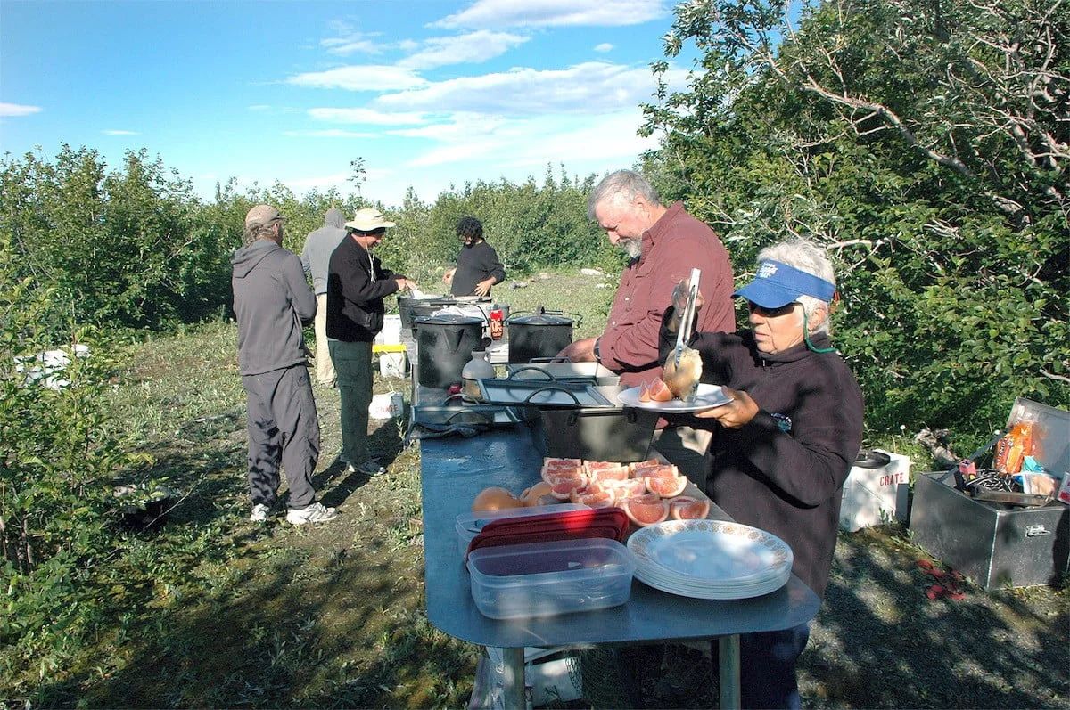 People preparing food outdoors near bushes under a blue sky; cooking station visible.