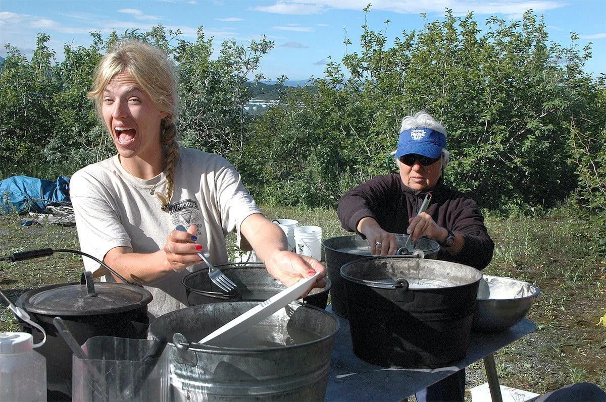 Two people cooking outdoors: one blonde, excited; one older, working over a pot.
