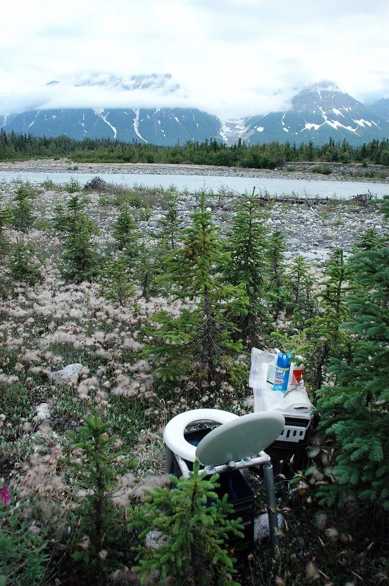 Toilet in a field with river and mountains in the background under cloudy skies.