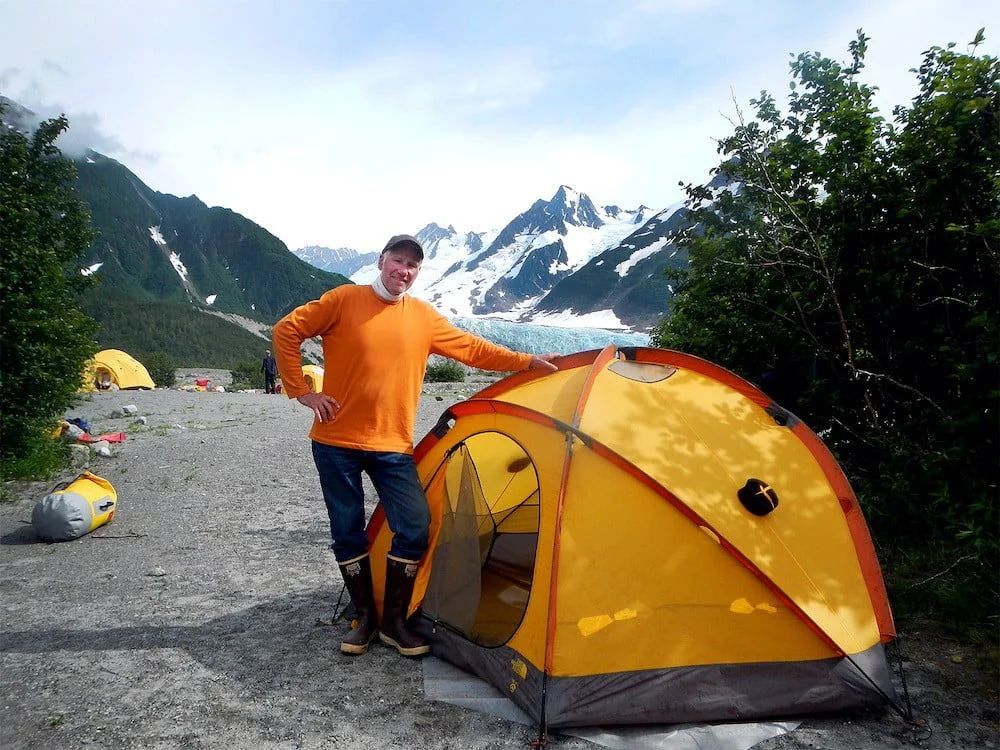 Man in orange shirt stands by a yellow tent, with mountains and glacier in the background.
