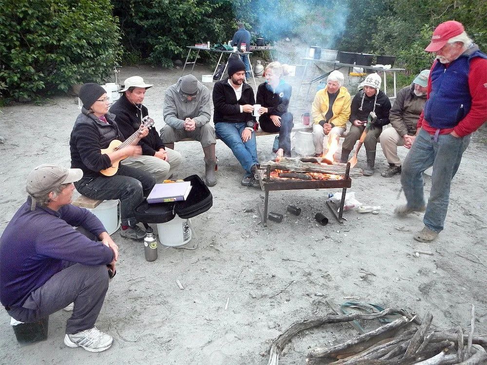 People around a campfire grilling food and playing music, outdoors.
