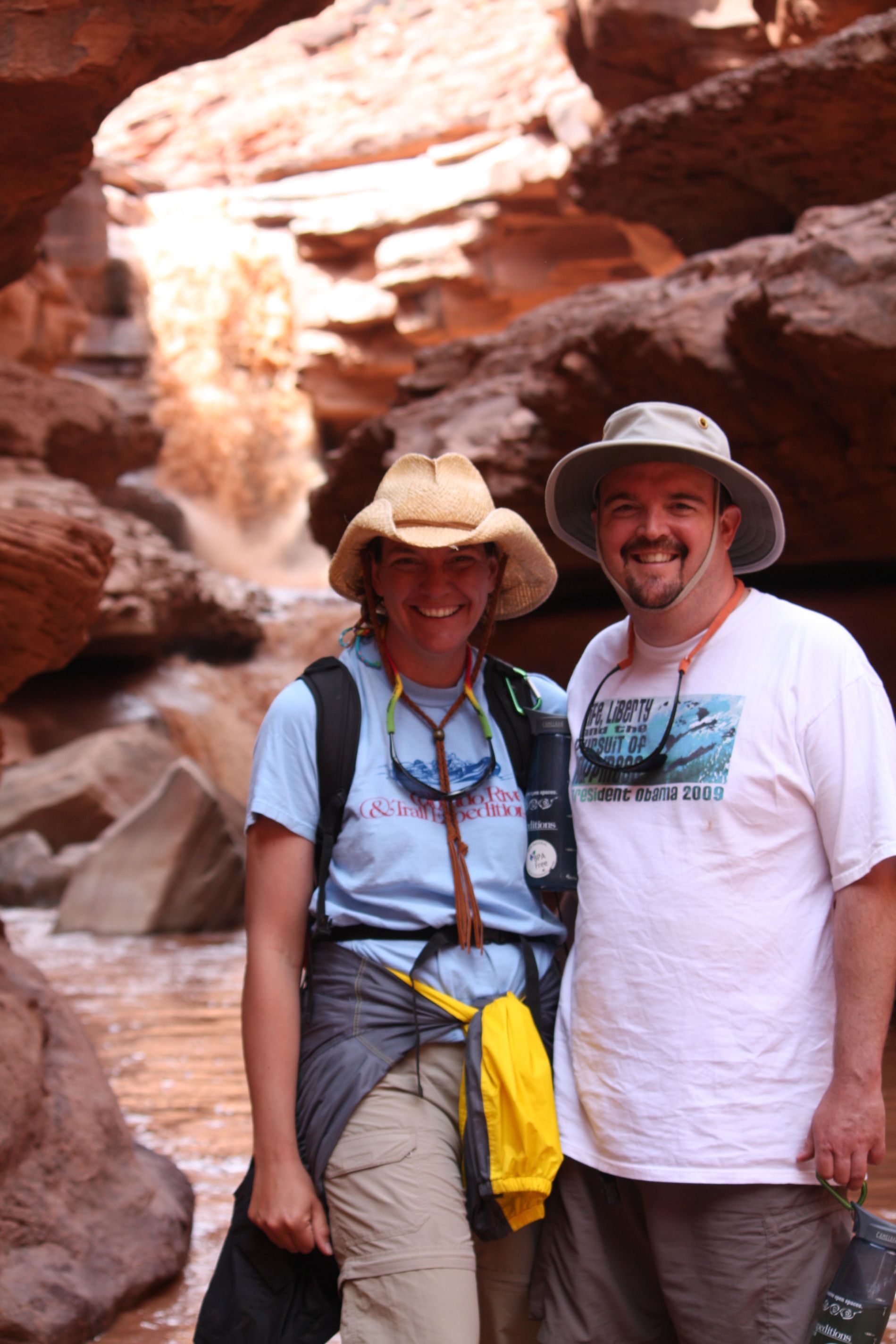Couple poses in a canyon, woman in a hat, man in a white tee, water cascading behind them.