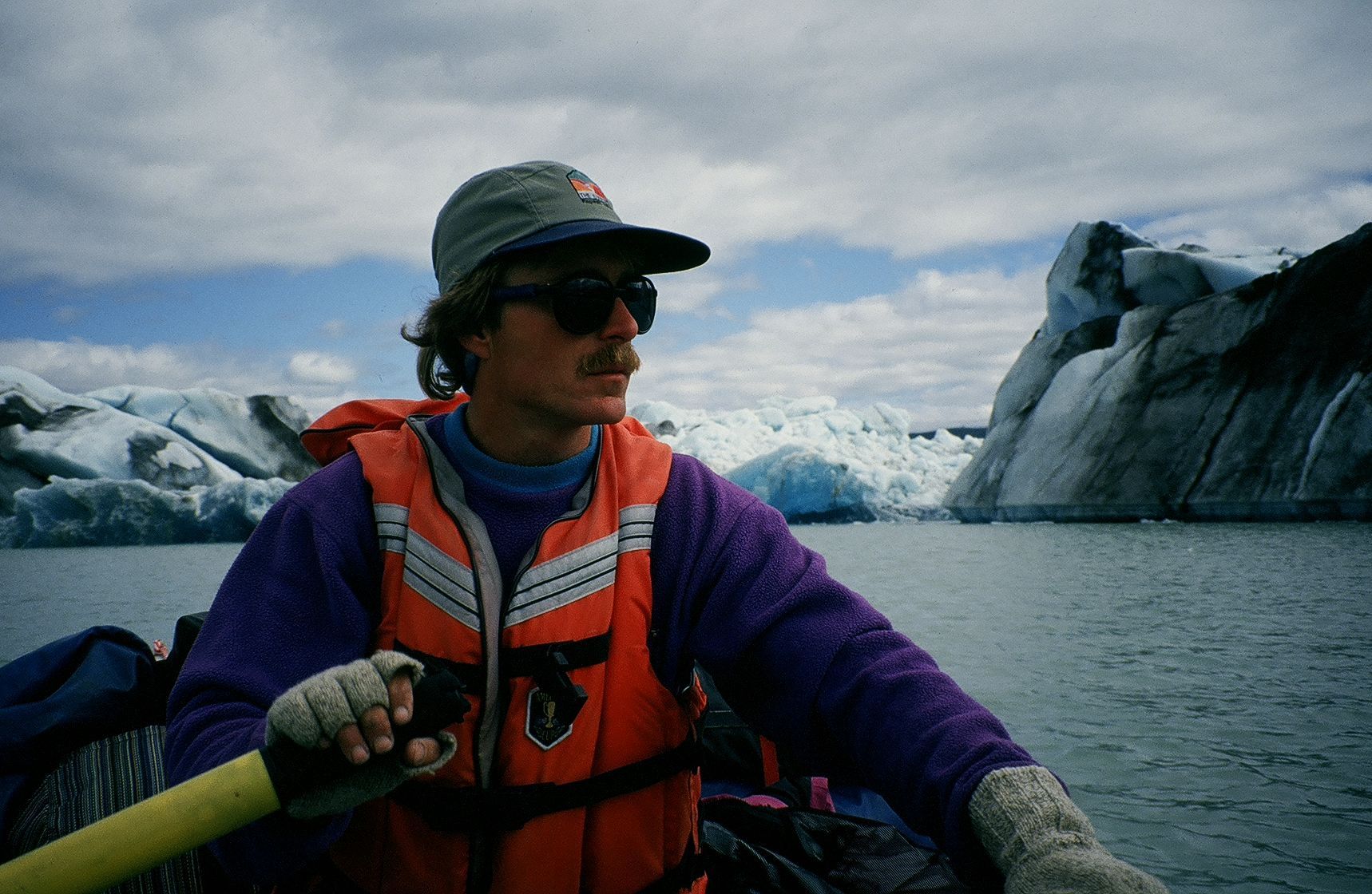 Man in a boat, paddling in front of a glacier, wearing a life vest and hat.