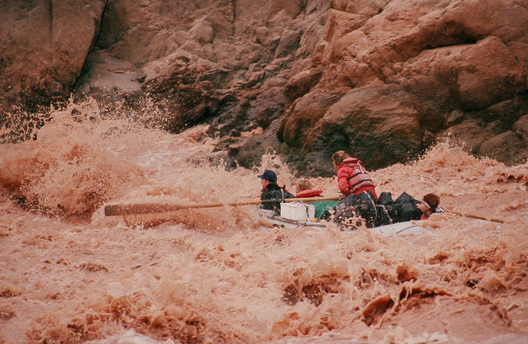 People in a raft navigating a turbulent, muddy river between reddish-brown rock formations.