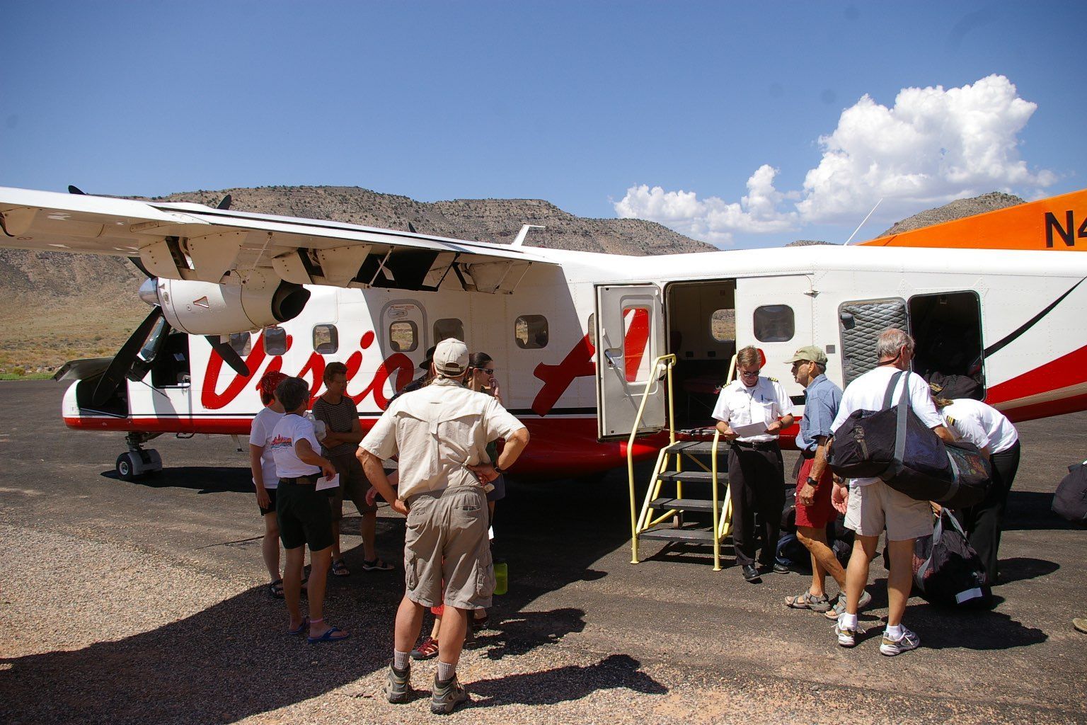 People boarding a white and red plane on a gravel runway with mountains in the background under a blue sky.