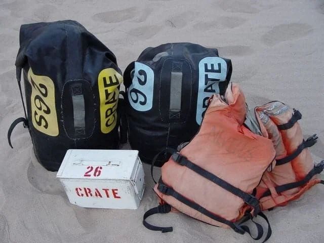 Two black backpacks and two orange life vests with a white crate, all on sand.