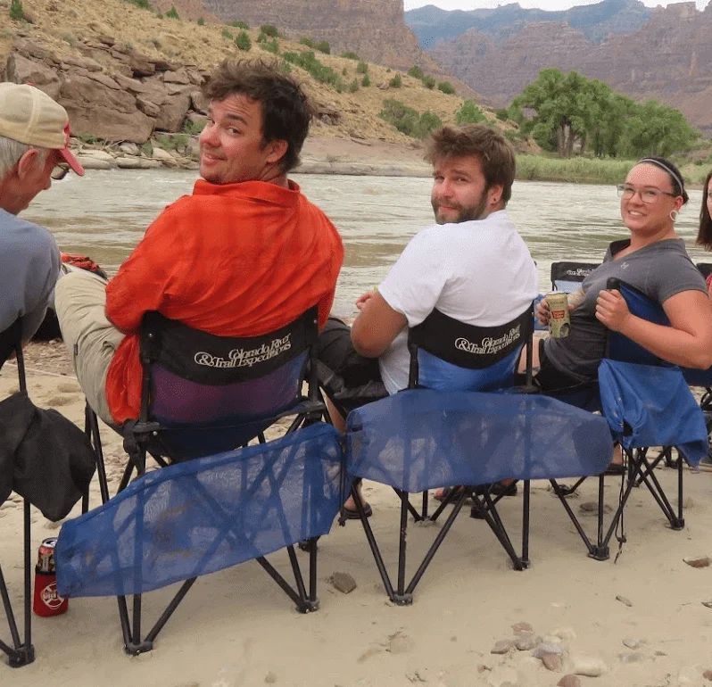 People seated in camp chairs by a river, looking towards the camera.