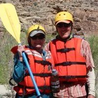 Two people in life vests and helmets smile while holding a paddle, near rocks.