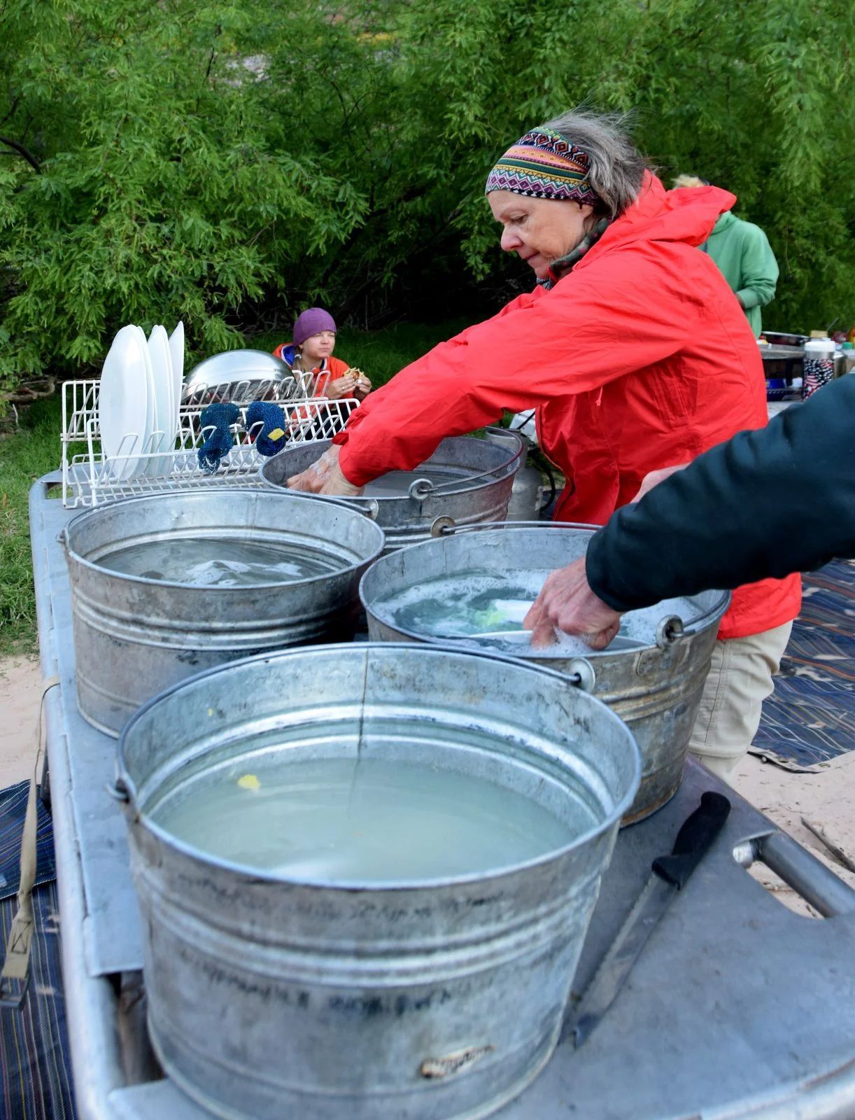 Woman in red jacket washing dishes in metal buckets outdoors.