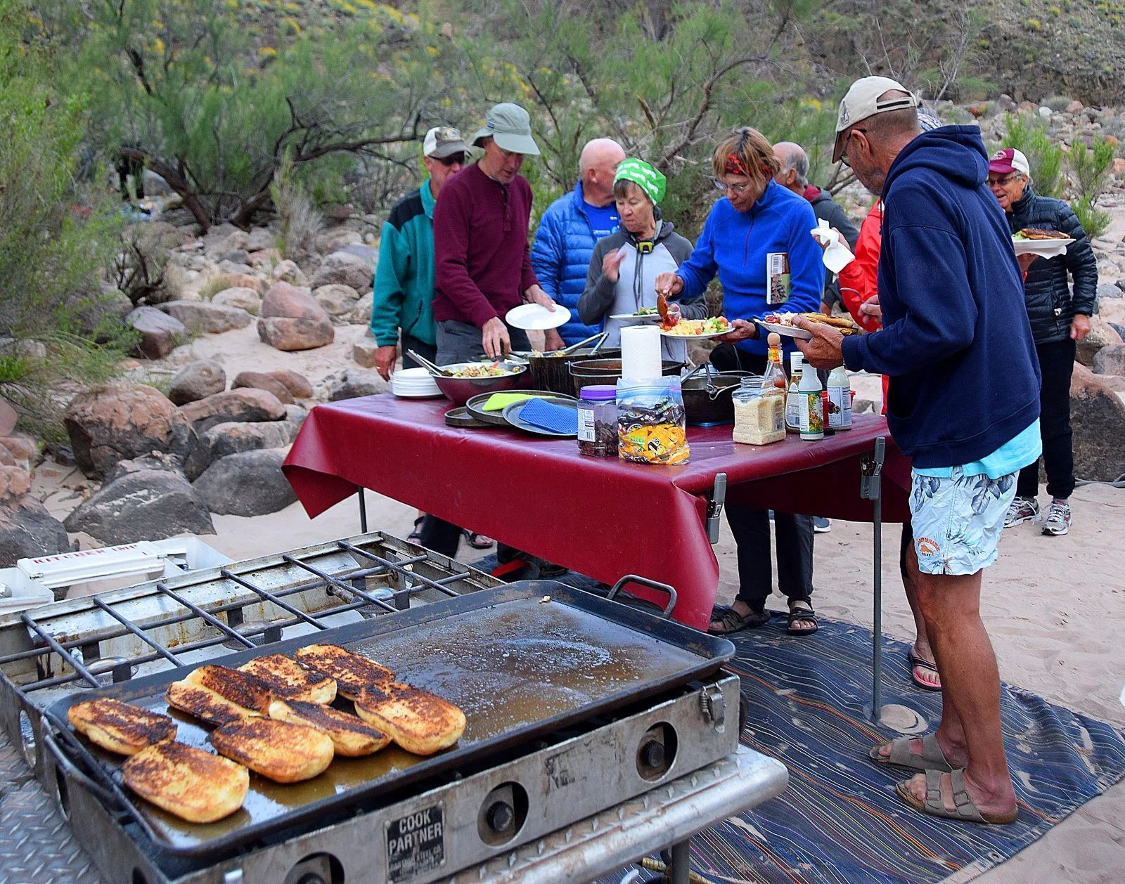 Group of people gathered around a table with food, cooking on a grill outdoors.