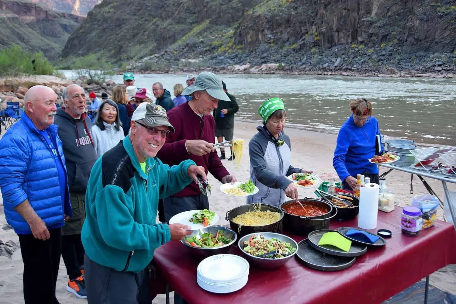 People serving themselves at a riverside buffet, mountains in background. Food includes salads and pasta dishes.
