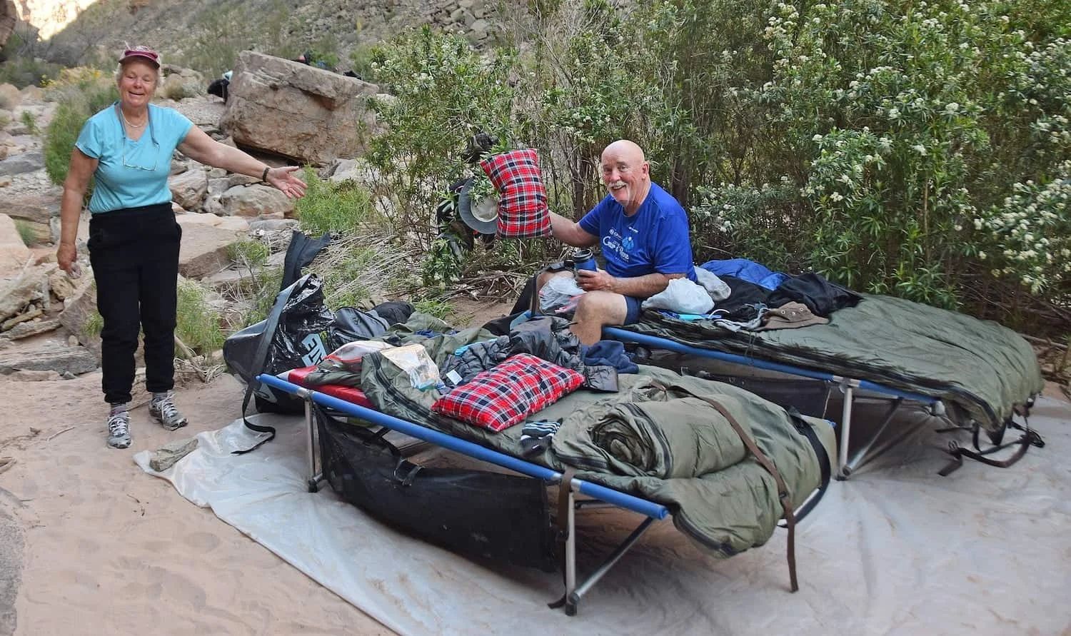 Two people by cots in a natural setting. One person points, the other holds a pillow. Green sleeping bags.