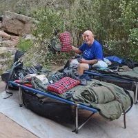 Man seated on cot, holding pillow. Camping gear visible; natural outdoor setting.