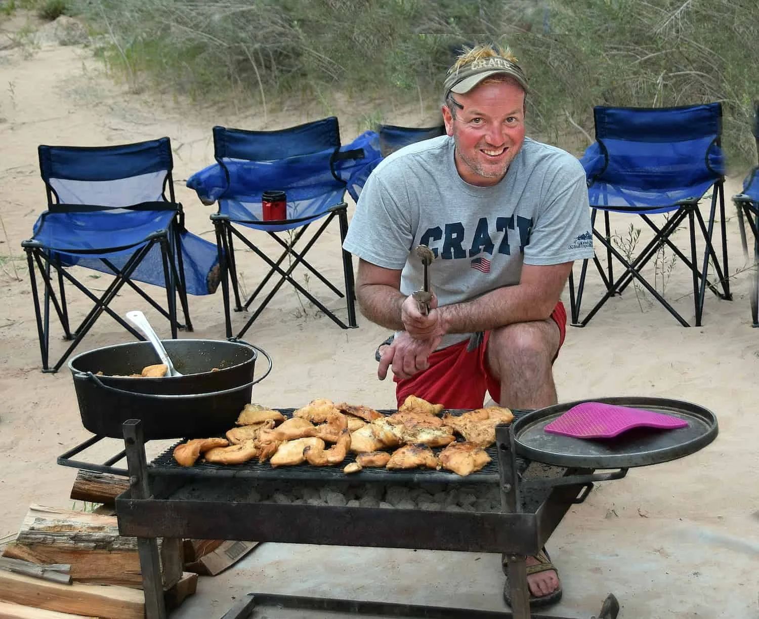 Man grilling food outdoors, smiling, with camping chairs and pot.