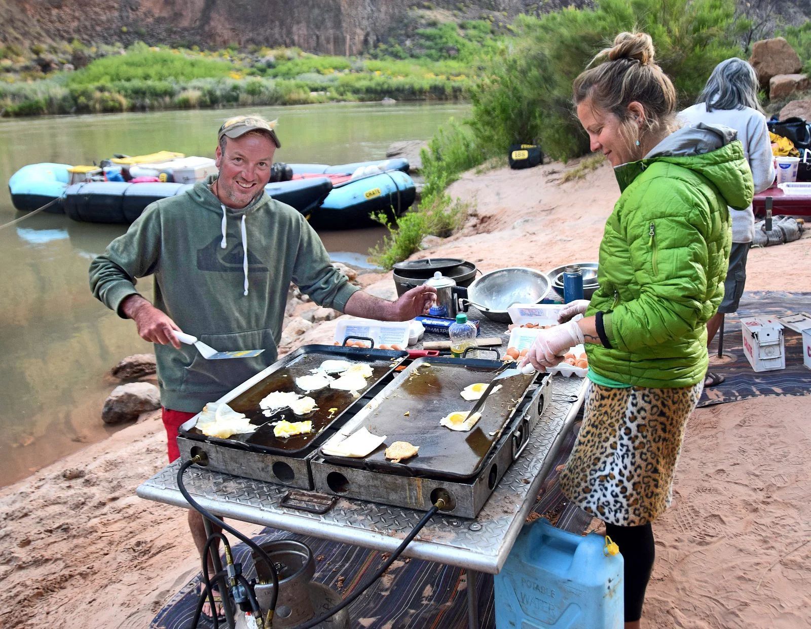 People cooking outdoors on griddles at a riverbank campsite. Man smiles while flipping food; woman pours ingredients.