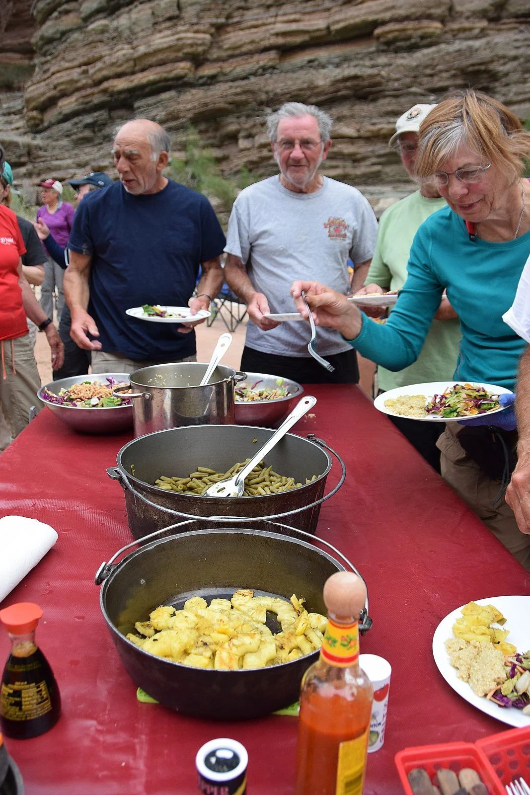 People serving food from pots on a red table outdoors. Several hold plates.