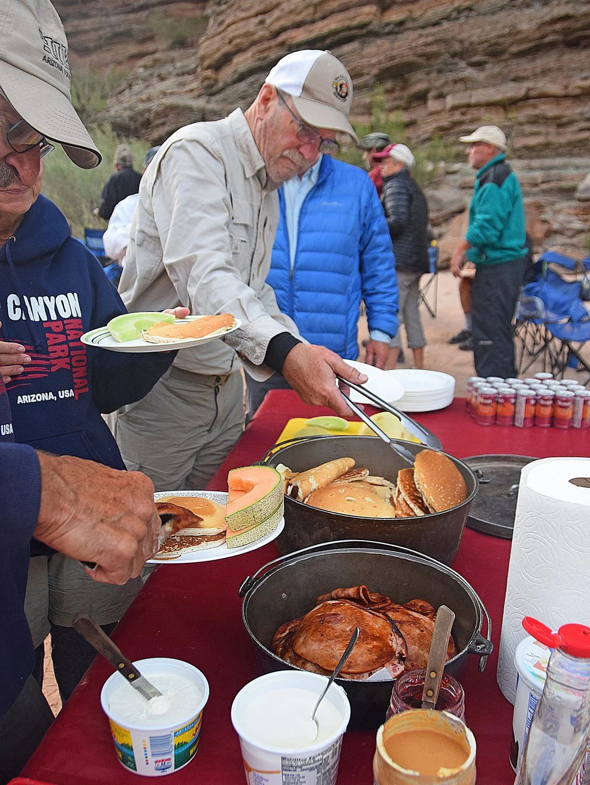 People at a picnic with food: fruit, bread, and yogurt. They are outdoors, near a canyon.