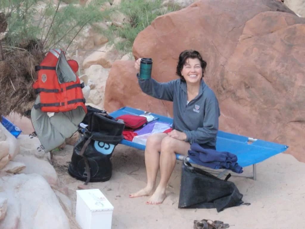Woman smiling, holding a mug, sitting on a camp cot by a rocky beach. Life vest and gear nearby.