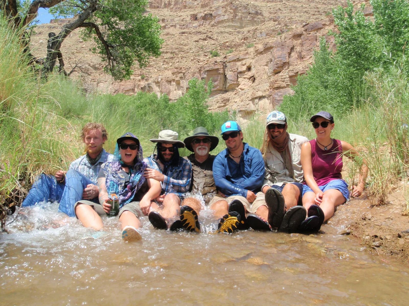 Group of people sit in a shallow stream, enjoying a sunny day outdoors.