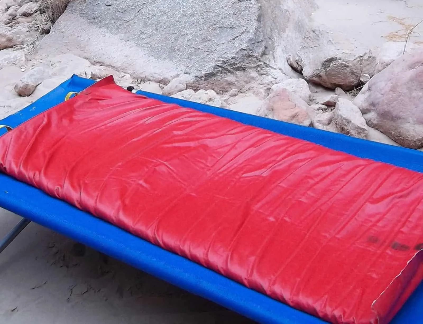 Red sleeping pad on a blue cot, near rocks and sand.