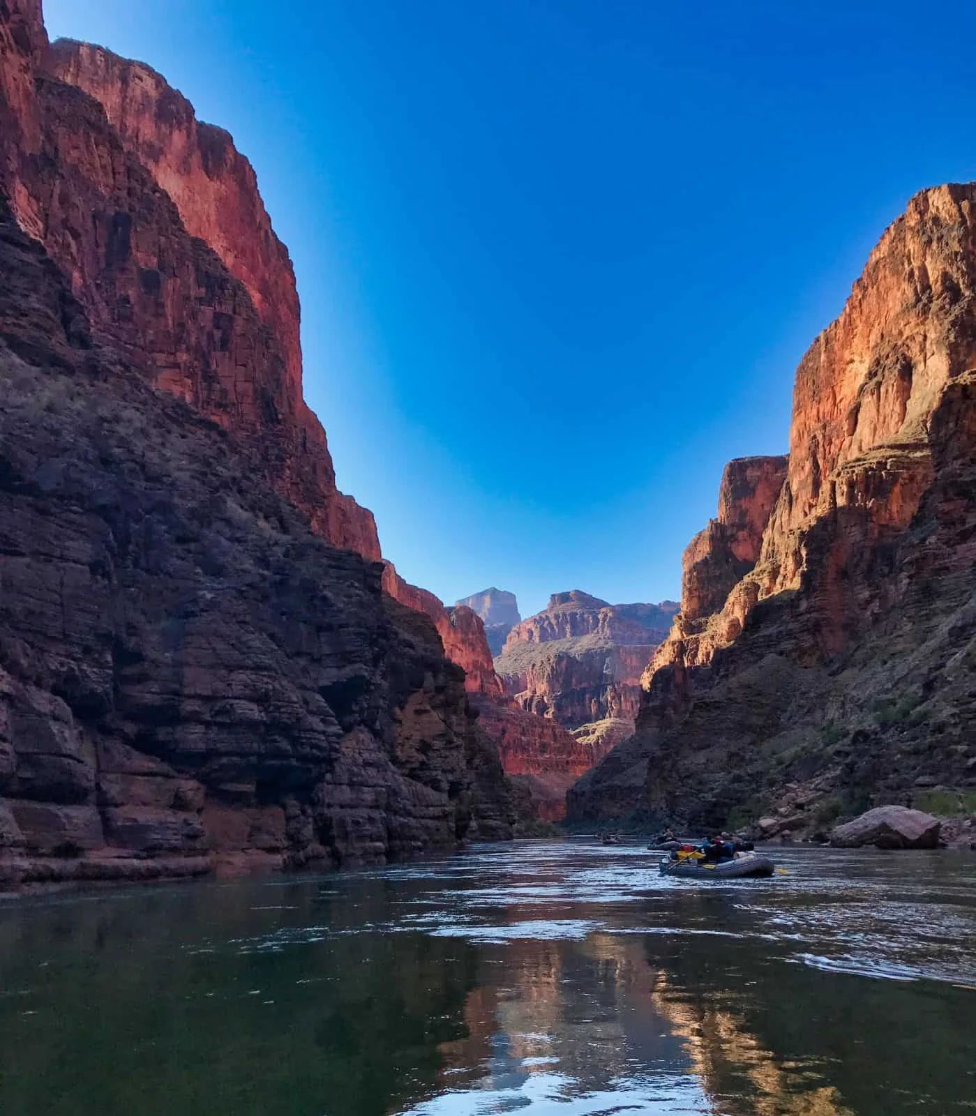 River flows through canyon, bright blue sky above. Sunlight highlights red rock walls; small boats on water.