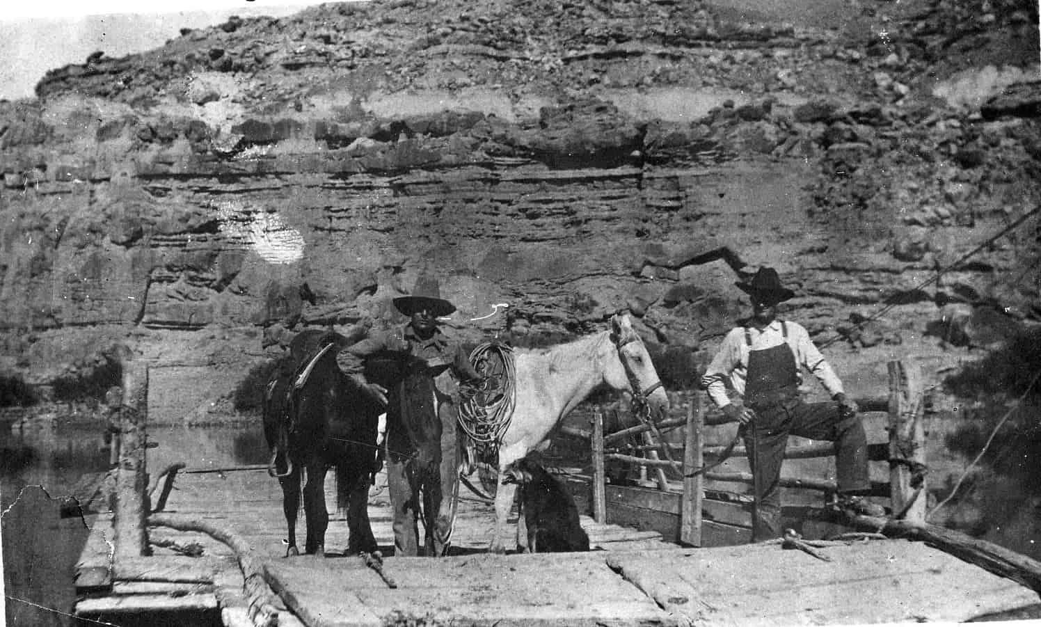 Two men with horses on a raft, near a rocky cliff.